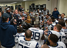 A man speaks to American football players in uniform in a locker room in front of a large trophy.