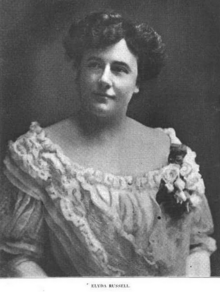 A white woman with dark hair in a bouffant updo, wearing a white frilly dress with a wide scooped neckline and a corsage