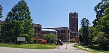 A red brick building is seen, with grass, trees, and construction equipment in the foreground. The building's tower and main entrance are to the right, with glass roofs and windows stretching to the left. Architecturally, the tower echoes the shape of the tower in the original building.