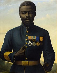 Portrait de face d'un soldat africain en uniforme avec quatre médailles. Sa main droite glissée dans son uniforme évoque la posture de l’empereur Napoléon.