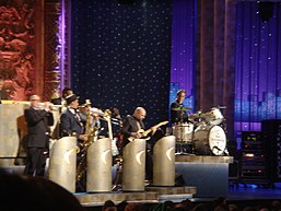 Several men are playing instruments, including trumpet, saxophone, trombone, and guitar, behind bandstands; at a higher elevation to the right of them is another man behind a drum kit and looking over at the band members; the bass drum is labeled 