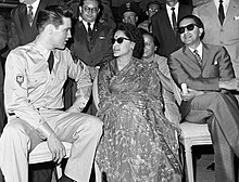 Photo en noir et blanc de trois personnes assises côte à côte : un jeune homme en uniforme militaire, en pleine conversation avec une femme en robe, et un homme en costume-cravate les bras croisés ; les deux derniers portent des lunettes de soleil