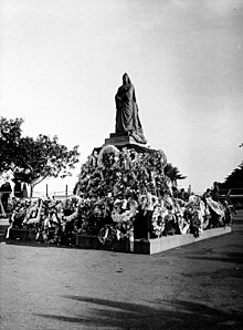 A black and white photograph of a statue of Queen Victoria, covered with dozens of flower wreaths.