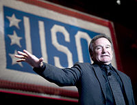 USOのロゴを背景に、黒字に細い白のストライプが入ったジャケット、紺色のシャツ、黒字に黒のチェック柄のネクタイを締めて、右手を広げながら喋るウィリアムズ