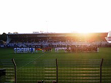 Dans un stade de football, avec le soleil couchant, deux équipes se tiennent alignées tenant un drapeau.