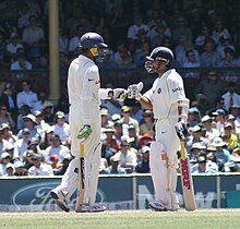 Two men wearing white uniforms shirts and trousers, blue helmets, gloves, pads and holding cricket bats meet one another on a cricket pitch to touch gloves. A crowd can be seen in the distance.
