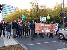 Des gens portant des banderoles et des drapeaux sont debout au milieu d'une rue, face aux voitures. À l'arrière-plan figure une rangée d'arbres et une tour de bureaux est située en haut à gauche de l'image