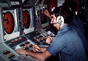 Un marin avec une chemise bleue est assis a une console qui possède un grand écran circulaire et des boutons de commande.