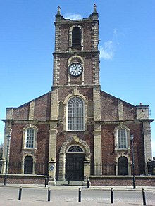 A brick church with stone dressings seen from the south. The west tower has a clock and pinnacles, and along the south face of the body of the church are Georgian-style windows.