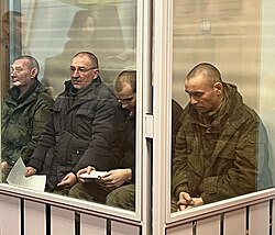 Four Russian soldiers, two head down, holding their prosecution documents, sit in a box with glass in the court room mid-session.