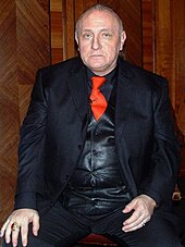 Photographie en couleurs de Richard Bandler, assis et face à l'objectif. Il porte un costume bleu et une cravate rouge. Il est chauve.