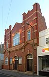 A tall building with deep red brickwork and yellowish stone dressings. There are two identical entrances: wooden doors in recessed stone doorways with prominent yellow keystones. A window to their left is in a similar recess. Two pediments flank a stone frieze with the words "THE SALVATION ARMY". The upper floor has two rectangular windows and a central arched window with another keystone.