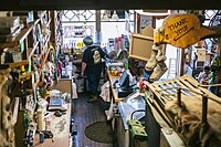 "Behind the counter with the cashier at Nagley's general store. It's an old and well-used building with creaky wooden floors and eroded countertops. There are bottles for flavored coffee, various boxes of hardware, a scale and price sticker wand, snacks, an antique cash register near a computer, an old dog sled, a pair of moccasins hanging from a sign that says Thank You, and hats that say Alaska. The front door is open.""