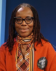 Portrait photographique de face d'une africaine d'une soixantaine d'années avec des deadlocks des lunettes et un percing dans la narine gauche.