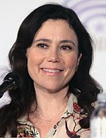 A woman with black hair tied back smiles while sitting behind a microphone.