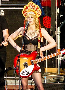 Woman with hands on hips, with a guitar, speaking into a microphone