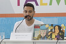 A man with short black hair and a white T-shirt sits at a table speaking into a microphone. Before him are two picture books on a display.