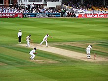 Мужчины в белых костюмах для крикета играют на зеленом поле для крикета посреди стадиона.