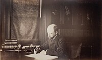 Henry Adams seated at desk in dark coat, writing, 1883