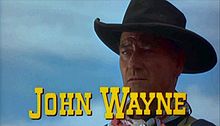 Image de film montrant en gros plan le visage d'un homme portant un chapeau de cow-boy, avec le texte « John Wayne » en lettres capitales jaunes en-dessous