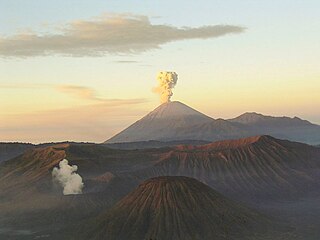 A brown volcano in the center with white smoke emanating from its peak, a cloudy sky fading from blue at the top through yellow in the middle to red at the horizon, and brown mountains in the foreground.