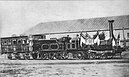 Auf einer alten Fotographie ist eine nach rechtss ausgerichtete Dampflokomotive mit zwei Wagons abgelichtet.