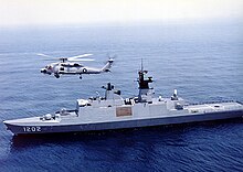Un navire de grande taille, gris, avec un hélicoptère volant au-dessus.