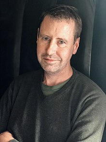 Portrait of author Tom Zoellner, wearing green sweater with arms crossed and in front of dark background.