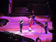 Four AC/DC members are shown from stage left; Johnson singing at front, Angus in red uniform with guitar, Stevie further right on guitar and Williams back left on guitar and singing.