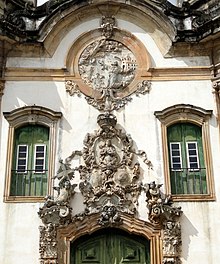 Photographie en couleurs. Détails de la façade, où le relief du portique, gris clair et très détaillé, est entouré de deux fenêtres et d'une porte vertes.