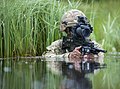 photographie en couleur montrant un soldat dans l’eau jusqu’aux aisselles qui tient un fusil d’assaut avec une imposante lunette montée dessus