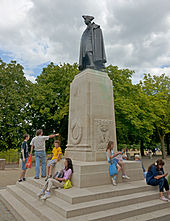 Темно-коричневая статуя мужчины в военной форме XVIII века, включая треугольную шляпу, на светло-коричневом каменном постаменте. Вокруг его базы разгуливают люди, одетые преимущественно в футболки и шорты. Есть деревья недалеко от