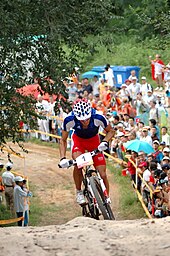 Homme debout sur les pédales d'un VTT portant une tenue bleu-blanc-rouge avec un public en arrière-plan.