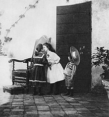 Photograph by Giuseppe Primoli: Two girls greet each other with a kiss; a boy looks on. Primoli, Giuseppe - Das Kusschen (Zeno Fotografie).jpg