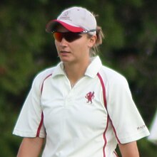 A woman wearing a white cricket t-shirt with red piping and a red dragon logo on the chest. She is wearing sunglasses and a grey and red cap, under which her light brown hair is up.