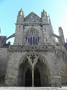 The West Porch with balcony and a small two gabled roof. The porch opening has a central colonnette with 2 arcs in "tiers-point à 3 lobes" and a central rosace of 5 lobes. A double door leads into the church, and in the trumeau is a 15th-century sculpture of the Virgin Mary with the child Jesus. In the tympanum are the arms of the Kermartins, the family of Saint Yves thought to date to the 13th/early 14th -century. Above the porch we see the first flying buttresses supporting the nave and a large elaborate window. Above this, a triangular gable has gothic pinnacles on either side.