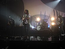 A performance shot of four men on stage. The man at left is turned away to his right, standing in a darkened area and holding a guitar almost vertically. The second man is singing into a microphone while playing a guitar. Behind and to his left is the drummer obscured by his kit. The last man is also shown in left profile and plays a guitar. Behind the performers is stage and lighting equipment.