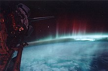 Abaixo, a Terra em branco e azul. Acima, erguem-se,flâmilas vermelhas da aurora em direção ao espaço. Parte do ônibus espacial é visível à esquerda.