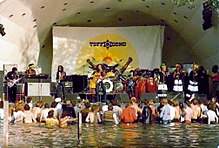 A मंच पर बैंड के प्रदर्शन को सुनते हुए, पानी में खड़े लोगों की भीड़