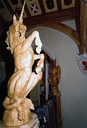 A photograph focusing on a large wooden sculpture of a unicorn, rearing on its hind legs. Behind it part of a wall and ceiling can be seen, the latter decorated with heraldic shields.