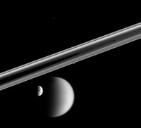 Circular complex rings of Saturn are seen at the low angle. The rings look like two grayish bands running parallel to each other from the left to right and connecting at the far right. Half illuminated Titan and Dione are visible slightly below the rings in the foreground. Two bright dots: one at the lower edge of rings and another above the rings can be seen. They are Prometheus and Telepso.