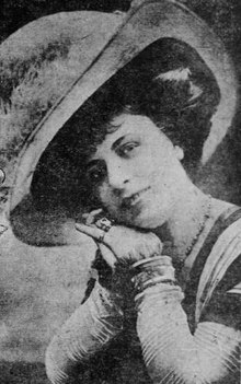 A young white woman wearing a large hat, smiling, her hands folded together and pressed against one cheek.