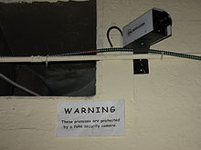 Camera on a concrete block wall, over a sign reading "WARNING: These premises are protected by a fake security camera"