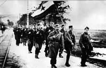 Black and white photo shows soldiers marching under the Yugoslav Partisan flag.