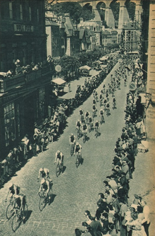 Photo en noir et blanc montrant un peloton de cycliste dans une rue avec de nombreux spectateurs sur les bords.