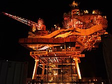 An offshore platform repurposed as an art installation, with walkways and foliage, illuminated by orange light at night.