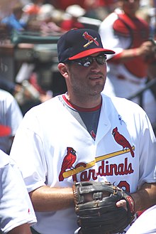 A white man standing still and wearing a white jersey. On the jersey is a yellow baseball bat with two red birds and "Cardinals" in red text under it. He is also wearing a black baseball cap with a red bird on a bat, sunglasses, and a black and brown baseball glove.