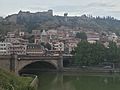 Pont en pierre de taille, dont deux des trois arches sont visibles, l'arche centrale couvre la rivière verdâtre (la Koura), celle de gauche une des berges sur laquelle circulent des automobiles, à l'arrière-plan un bâti dense inclut des immeubles de teintes variées : blanc, rose, gris devant des murailles de teinte grisâtre.