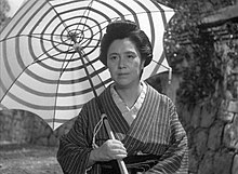 Hie rist eine schwarz-weiß Fotografie im Querformat zu sehen. Urabe Kumeko steht in der Bildmitte und hält einen riesigen Sonnenschirm der ein Spiralenmuster hat. Sie trägt gestreifte Kleidung.