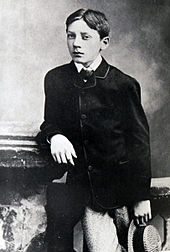 Photo noir et blanc. Garçon, habillé d'une veste, accoudé à une table et tenant un chapeau, regardant à gauche.